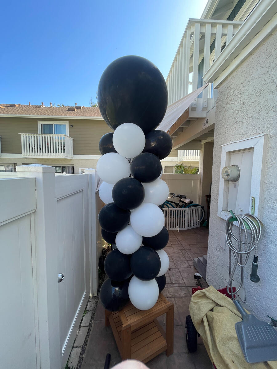 Tall black-and-white balloon column with a large black topper, resting on a wooden stool on a patio.