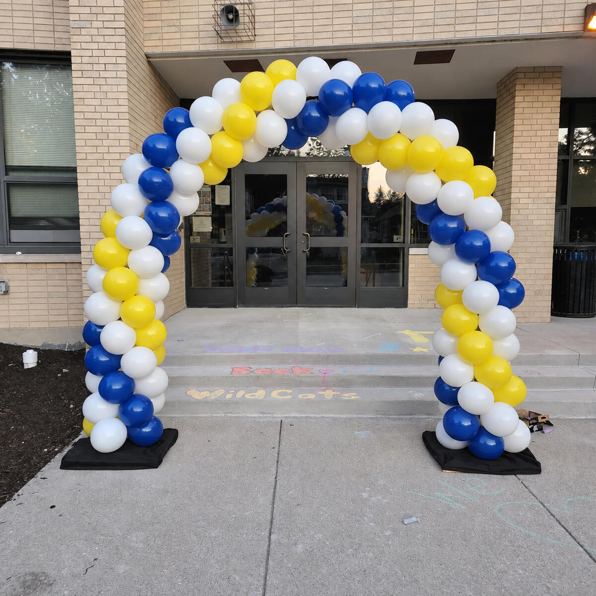 Blue, yellow and white balloon arch framing a building entrance, with both floor bases visible.