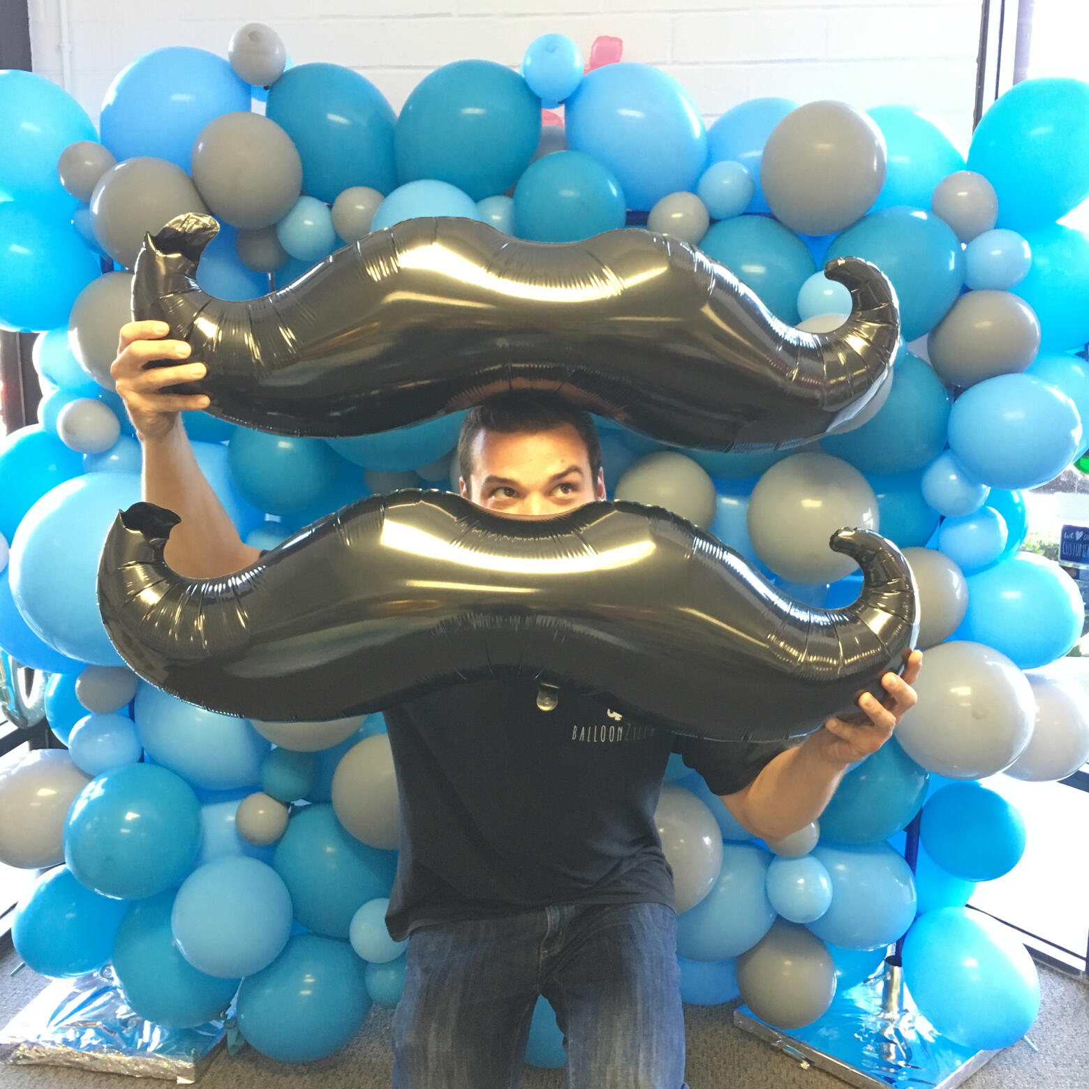 Josh holding two black moustache-shaped balloons in front of a blue-and-gray balloon wall.